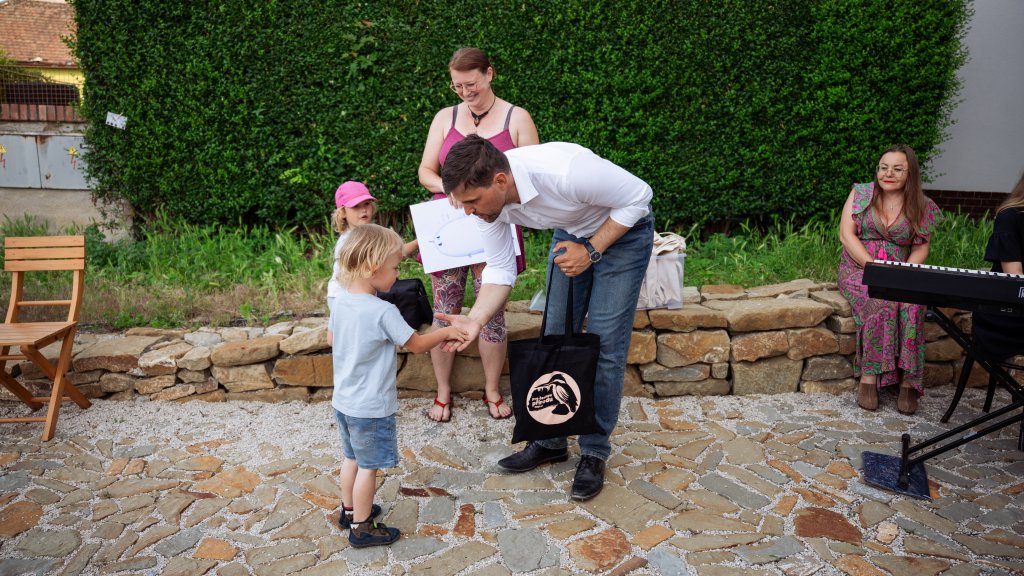 A man in a white shirt bends down to shake hands with a young child during an outdoor community event.