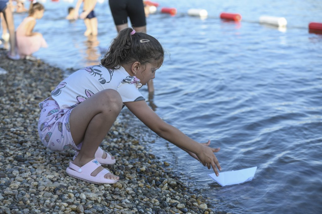 A young girl crouches by the water’s edge on a pebbled beach, gently placing a small paper boat into the water.