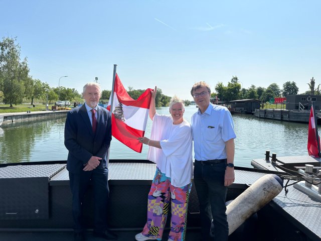 Three people with Austrian Flag stand in front of Danube River. 