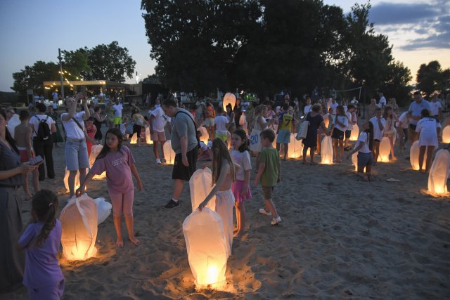 People of all ages gather on a sandy beach at dusk, holding glowing paper lanterns as they prepare to release them into the evening sky.