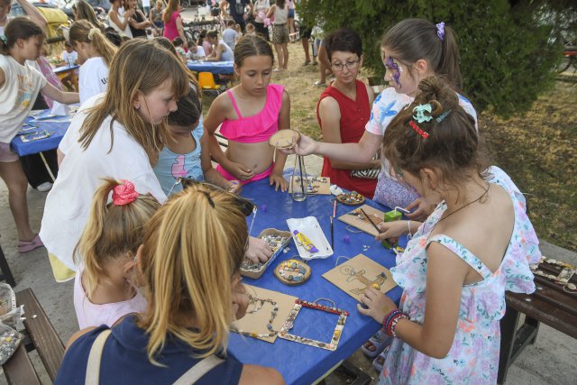 A group of children gather around a table at an outdoor craft workshop, creating art projects with wooden pieces and colourful decorations.