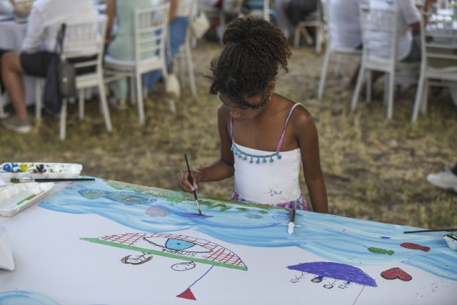 A young girl paints a colourful scene with umbrellas and water on a large sheet of paper during an outdoor art activity.