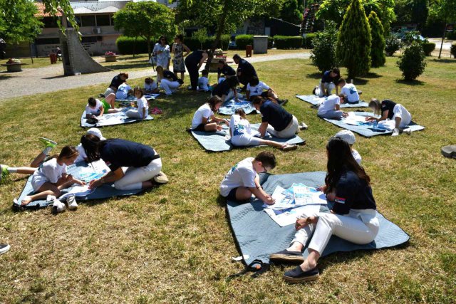 Children sit on mats outdoors, drawing and painting with the help of adults during an art activity on a sunny day.