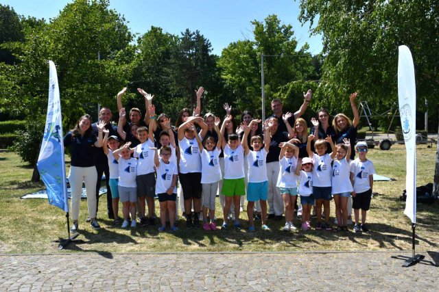 A group of children and adults wearing matching white shirts pose outdoors with their arms raised in celebration between two event banners on a sunny day.