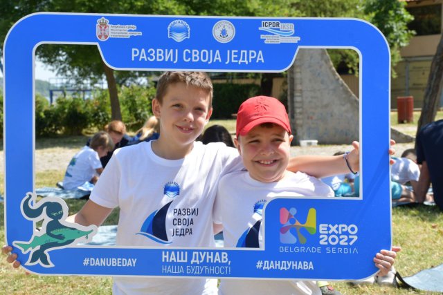 Two smiling children wearing white event shirts pose together outdoors, holding a large blue photo frame with logos and Serbian text promoting the Danube and EXPO 2027.