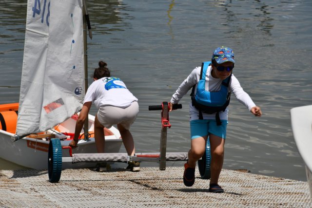 Two young sailors prepare their small sailboat at the water’s edge; one adjusts the boat on a dolly while the other runs ahead in sailing gear and a life jacket.