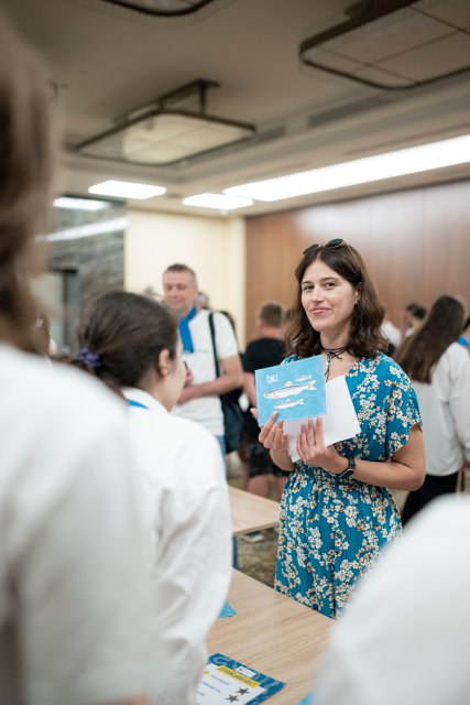 A woman in a blue floral dress holds up a booklet and speaks to a group of students during an indoor educational Danube Day event.