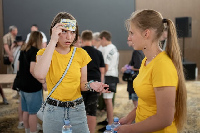 Two young women wearing matching yellow shirts play a "what am I?" guessing game while standing at a table with water bottles during an indoor Danube Day event.