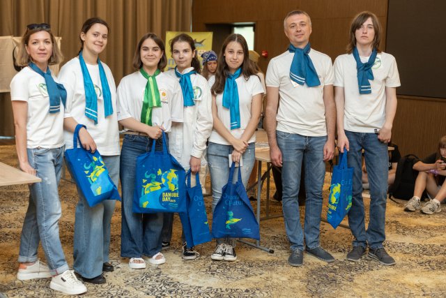 A group of seven people wearing white shirts and blue or green scarves pose together indoors, holding blue Danube Day tote bags after an event.
